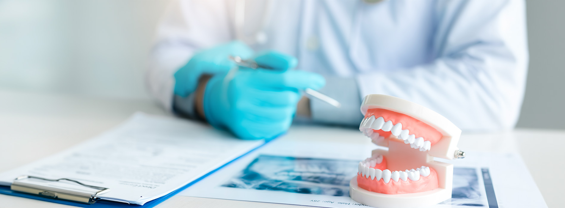 The image depicts a dental professional, likely a dentist or dental hygienist, seated at a desk with a tray of dental instruments and a mouth model, wearing gloves and focusing on paperwork.