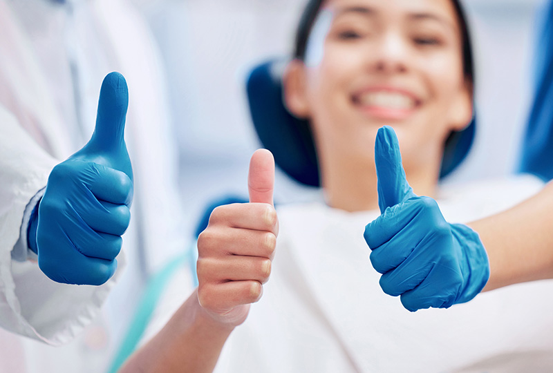 Image  Two thumbs up with blue medical gloves, a smiling person in the background wearing medical attire.
