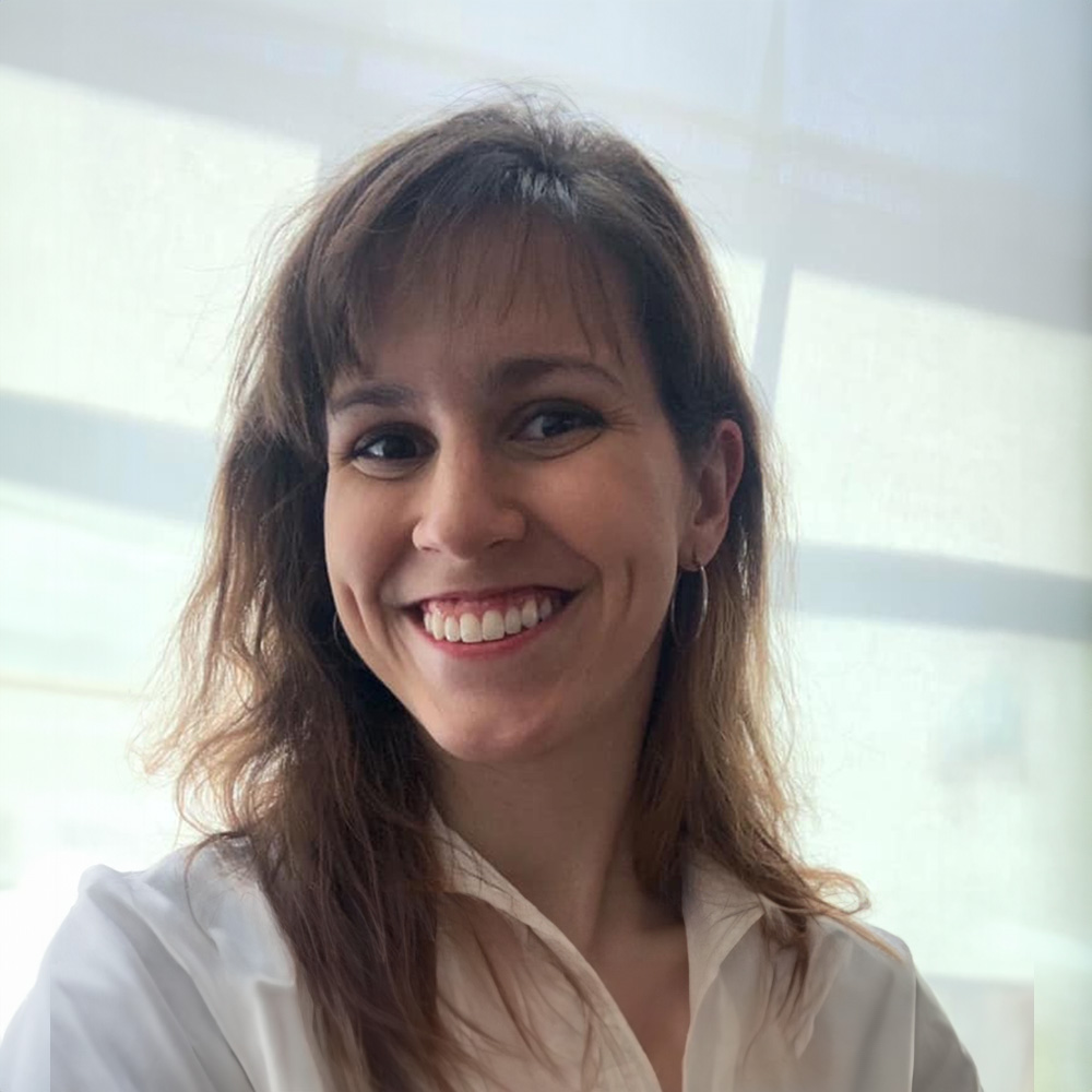 The image shows a woman with a smile, wearing a white shirt and sitting in front of a window.