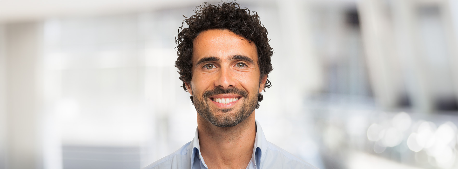 An adult male with curly hair, wearing a blue shirt and a lanyard, smiling at the camera in an indoor setting.