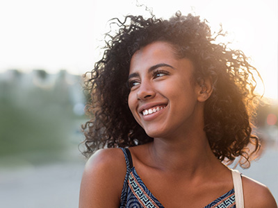 The image is a photograph of a young woman with curly hair, smiling at the camera. She has dark skin and is wearing a sleeveless top with a patterned design. The background is blurred but suggests an outdoor setting during daylight hours.