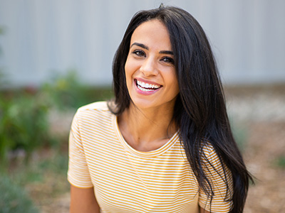 A young woman with long dark hair is smiling, wearing a yellow top and standing outdoors.