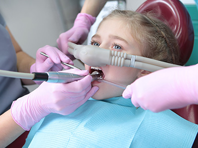 A young girl receiving dental treatment in a dental chair, with a dentist and dental hygienist working on her teeth.