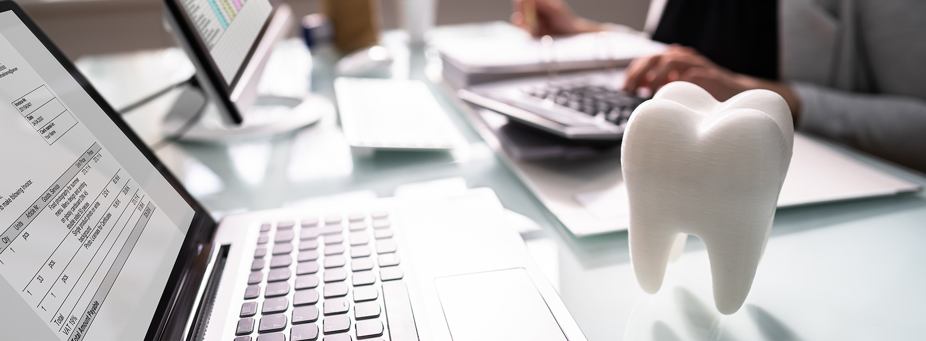 The image features a desk with various items including a laptop, a computer mouse, a keyboard, and some paperwork. There is also a model of a tooth placed on the desk. A person is partially visible at the top left corner of the image, working at the desk.