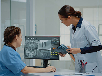 The image depicts a medical professional in a white coat standing next to a woman who is seated at a computer screen displaying an X-ray.