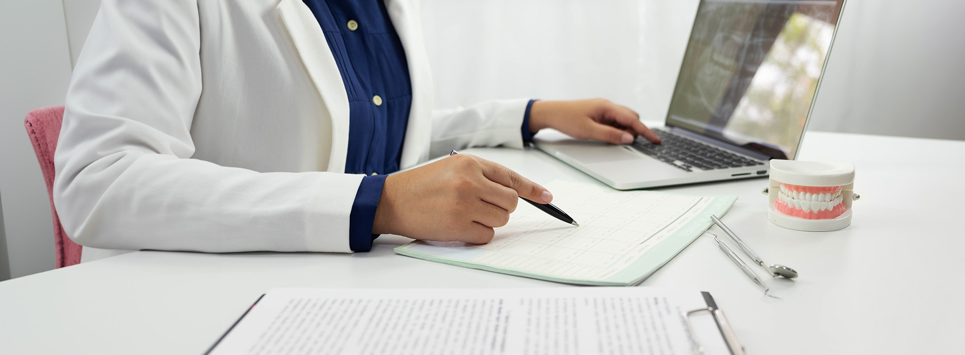 A person sitting at a desk with a laptop, papers, and pens, wearing a white lab coat.