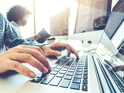 The image depicts a person typing on a laptop computer with multiple screens in the background, suggesting a professional or work environment.