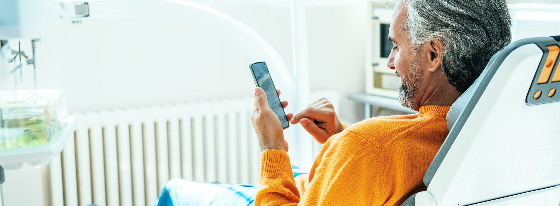 An elderly man in a medical setting, sitting in a chair and using his phone.