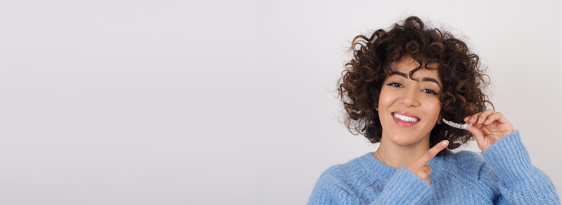 The image shows a person with short hair smiling at the camera, wearing a blue sweater and holding what appears to be a smartphone in their right hand.