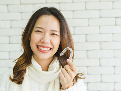 A woman with a radiant smile is holding up a dental retainer, showcasing it against her teeth.